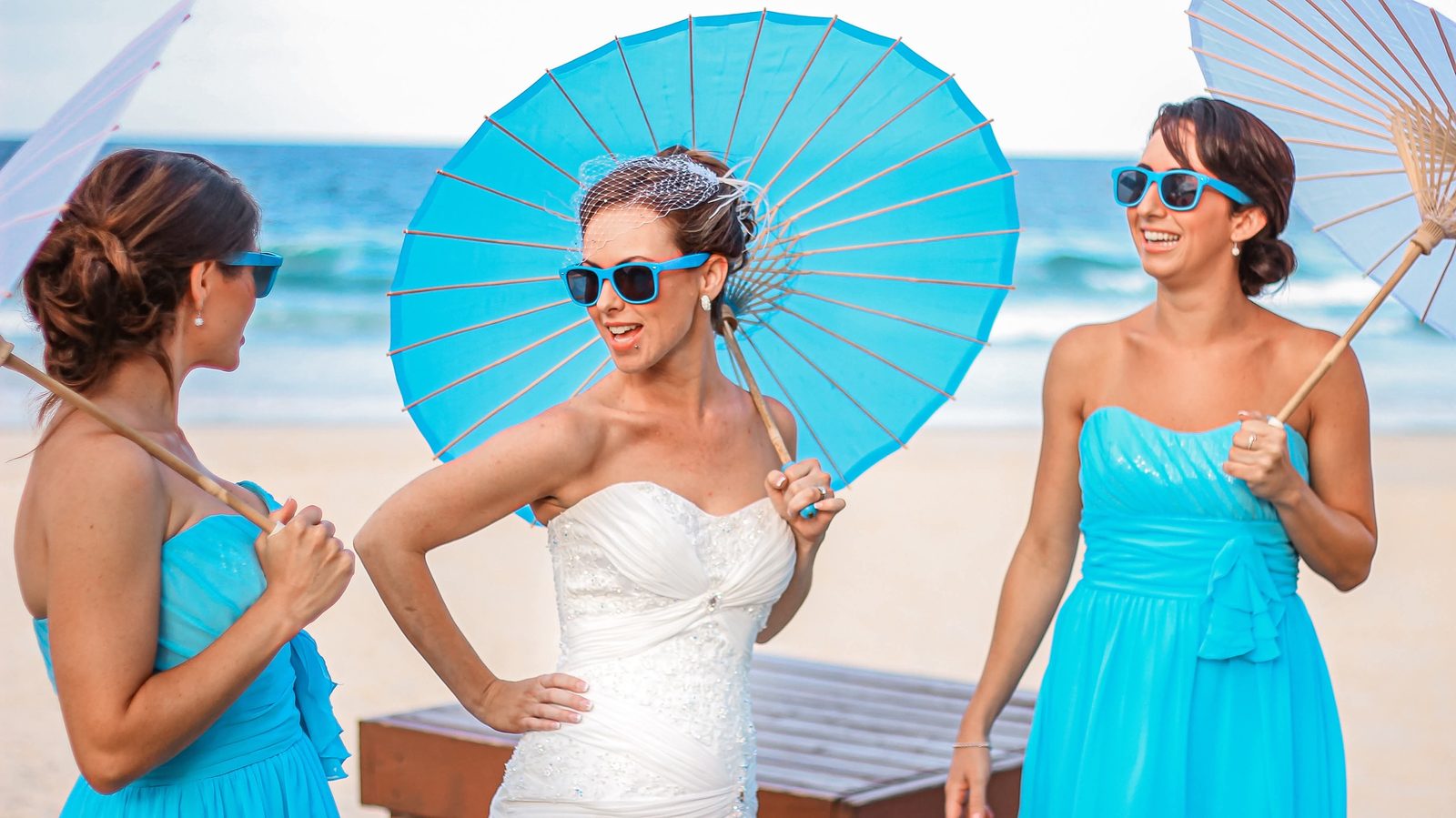 A bride laughing with her bridesmaids, all holding parasols, beside the sea