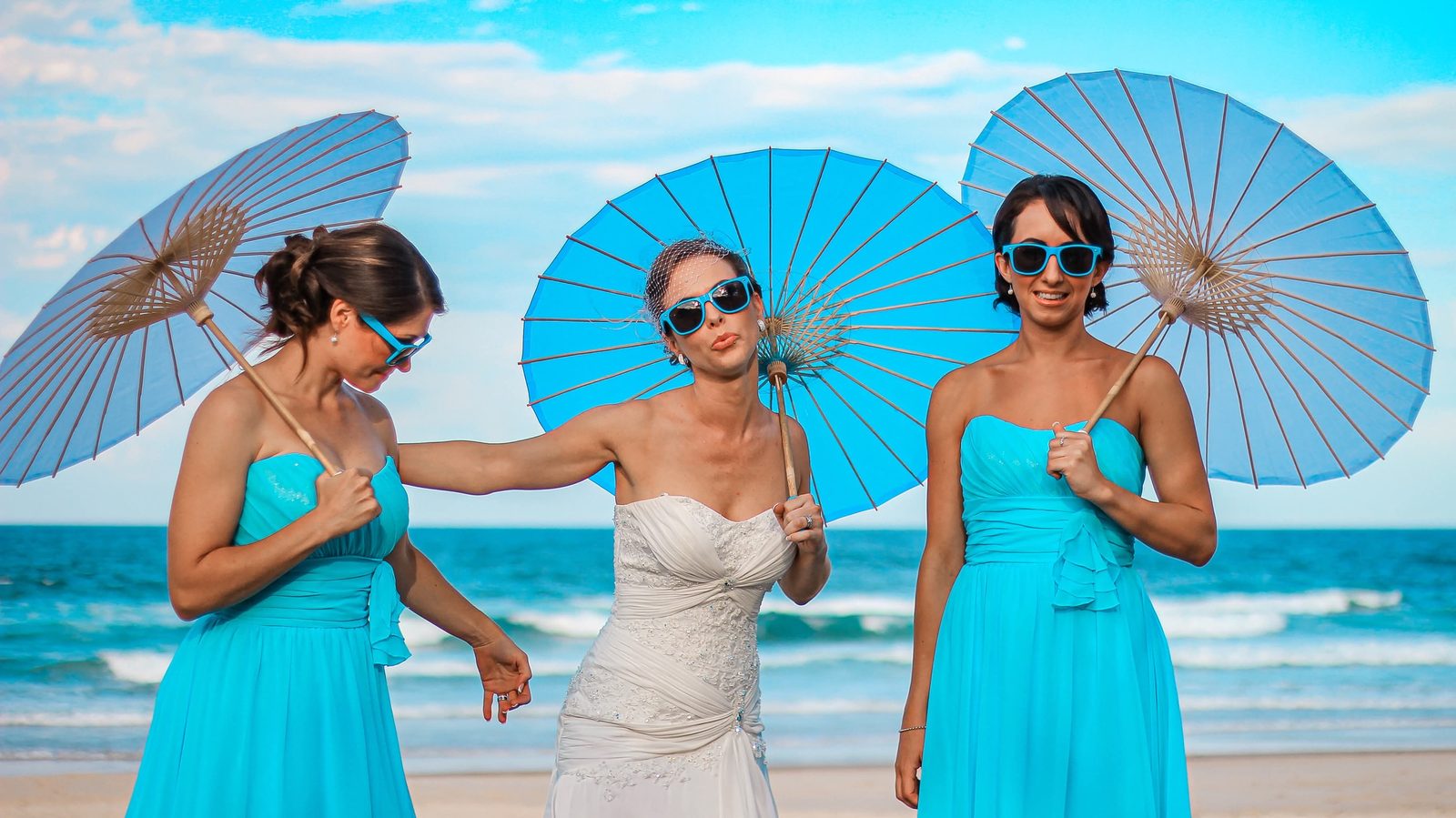 A bride and her bridesmaids with blue parasols and sunglasses on the sand