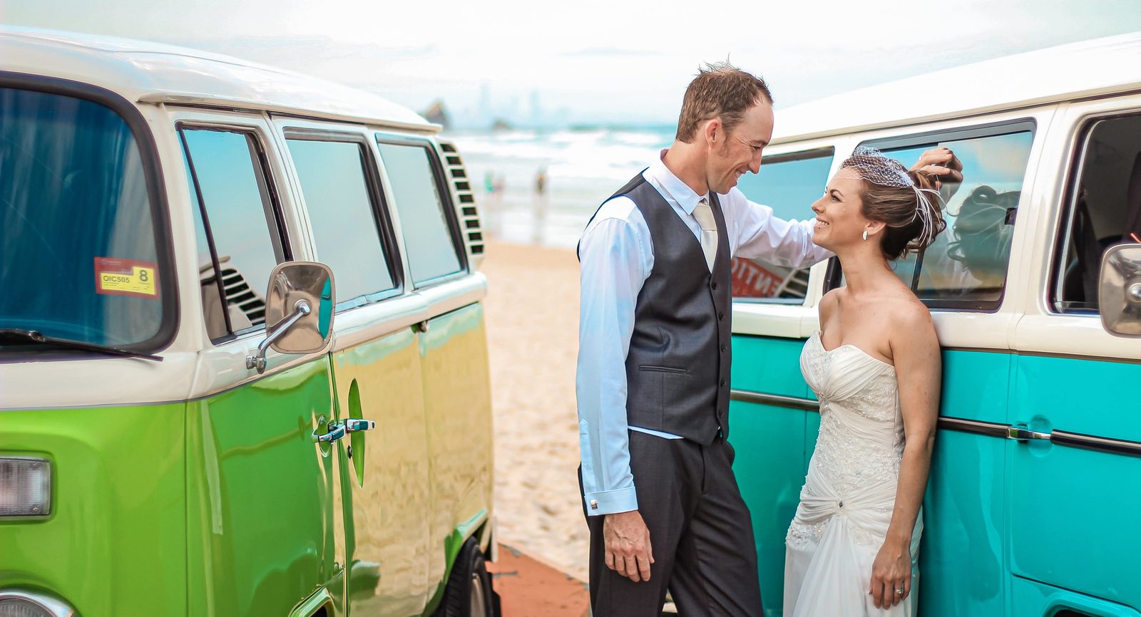 A couple smiling at one another between two vintage Kombi vans on the beach
