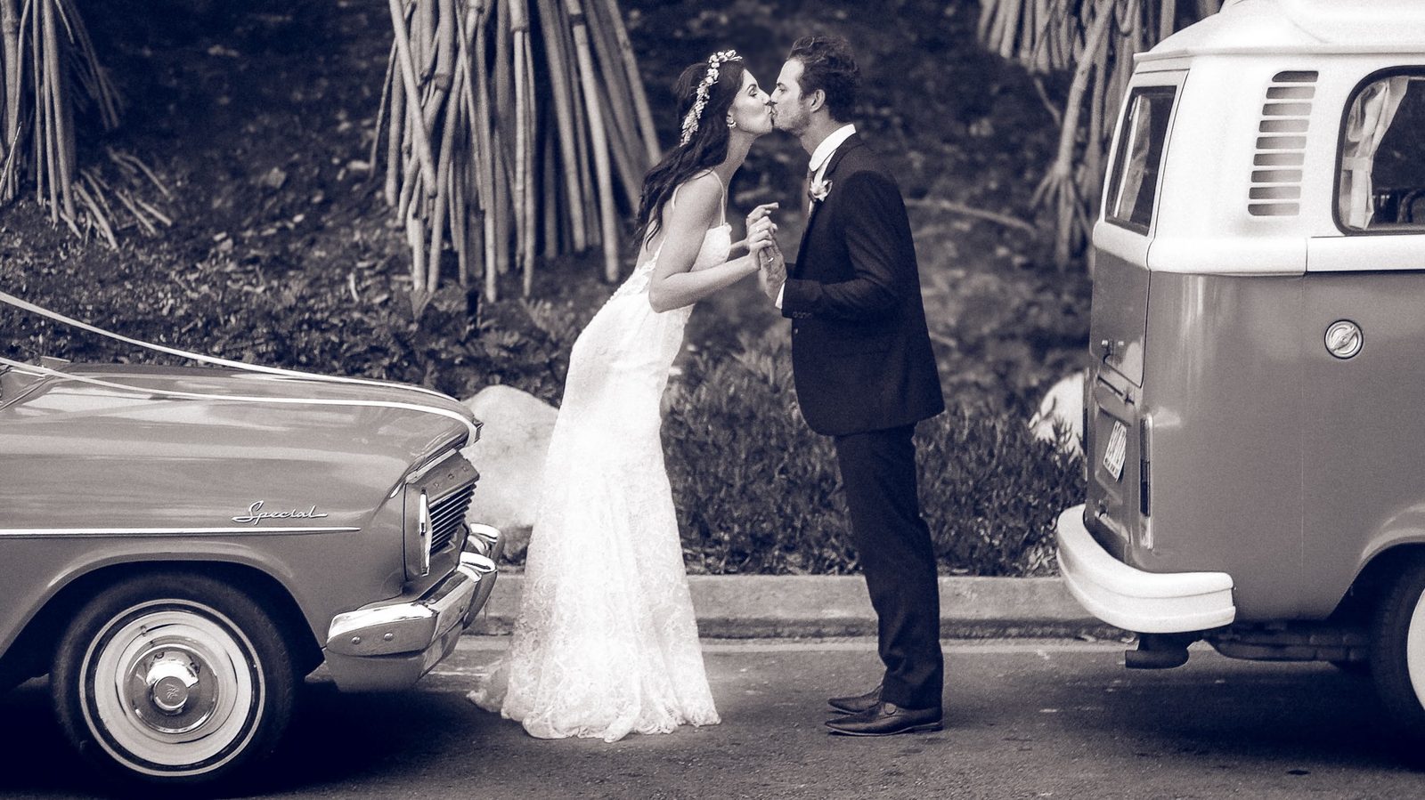 Black and white photograph of a couple kissing between a vintage car and a Kombi van