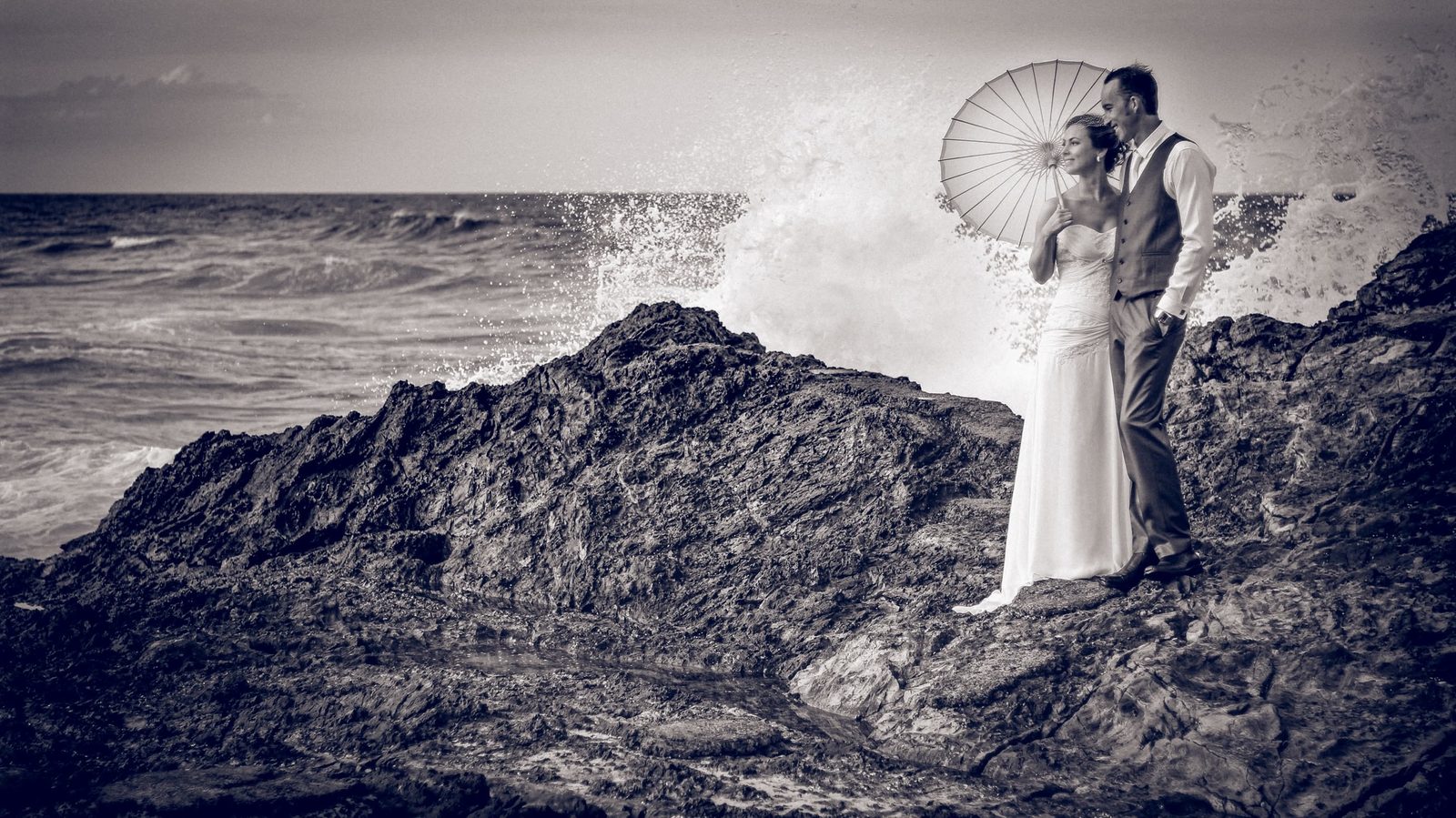 A couple sheltering under a parasol on rocks as a wave breaks behind them