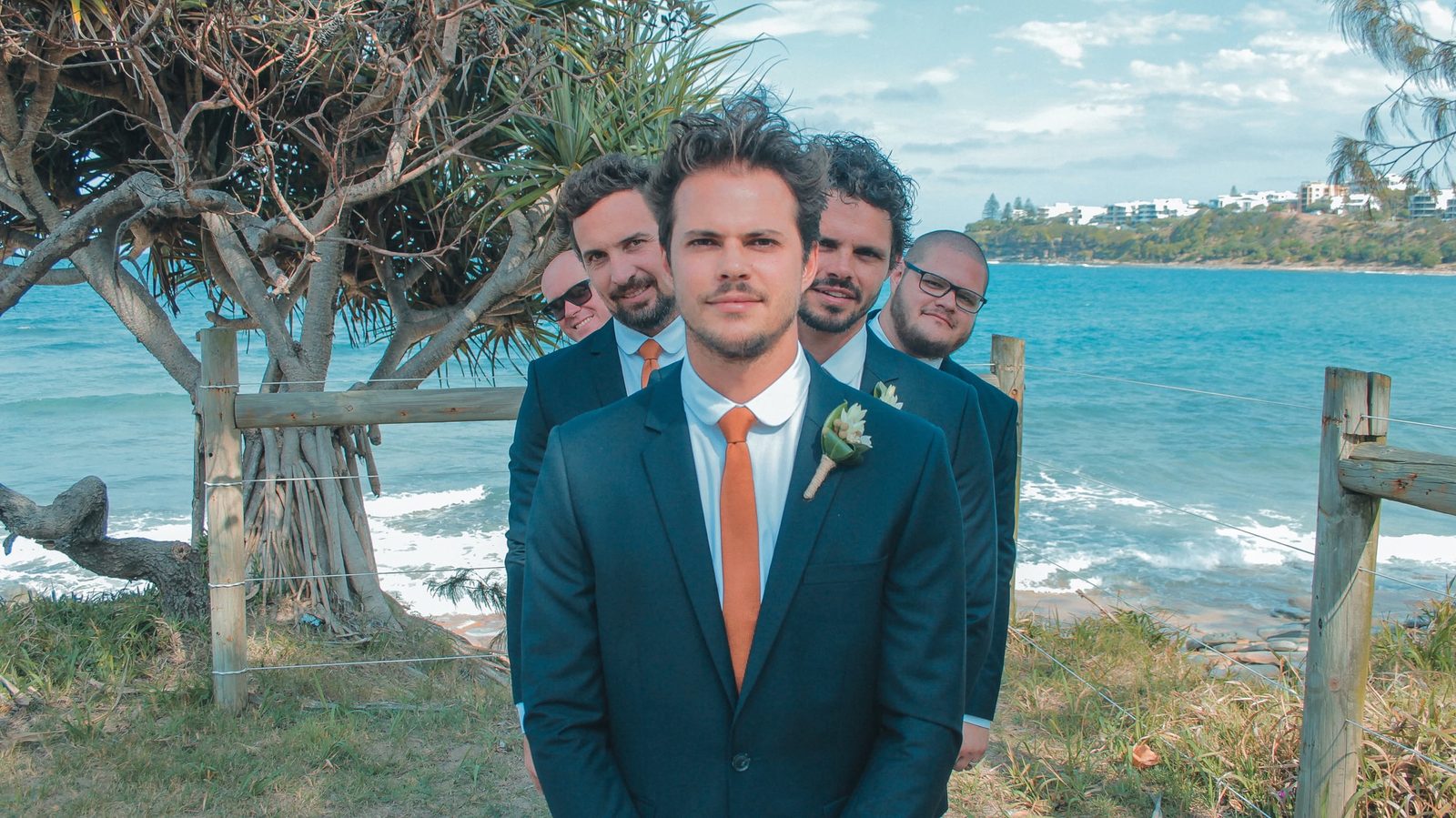 A groom and his groomsmen in navy suits and burnt-orange ties on a coastal headland
