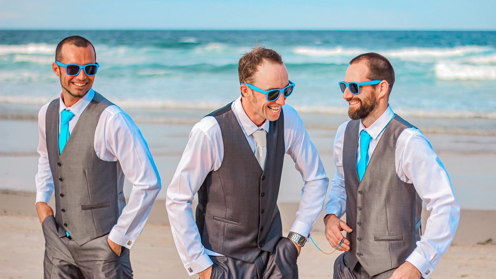 Groomsmen in grey waistcoats and bright blue sunglasses laughing on the beach